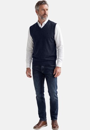 Uomo con capelli grigi e barba che indossa un maglione senza maniche blu navy sopra una camicia bianca, jeans scuri e scarpe di cuoio marrone, in piedi e che guarda a destra.