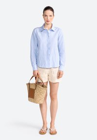 Lichtblauw gestreepte overhemden met lange mouwen, beige gehaakte shorts en een rieten tote bag met leren accenten. Beige slippers.