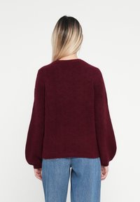 Burgundy színű kötött pulóver, kerek nyakkivágással és ballonujjakkal, puha textúrával és bordázott alsó résszel. Világos kék farmerrel párosítva.
