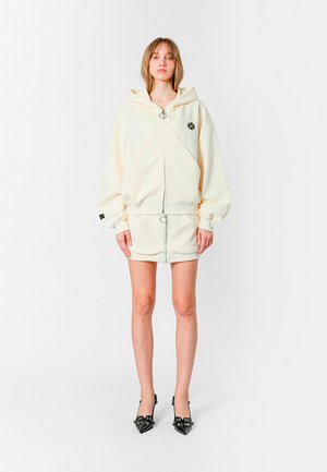 Sudadera color crema con corte oversized, capucha con cordones y cremallera frontal. Combinada con una mini falda a juego y tacones negros con punta.