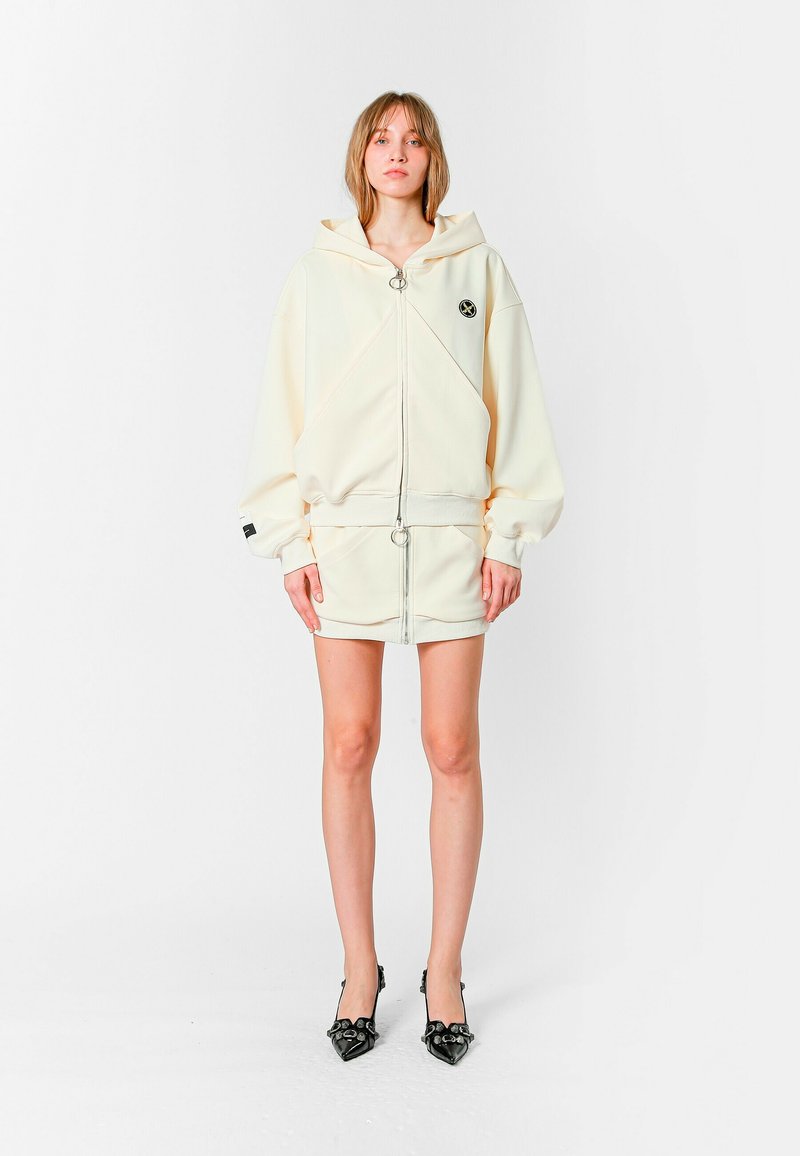 Sudadera color crema con corte oversized, capucha con cordones y cremallera frontal. Combinada con una mini falda a juego y tacones negros con punta.