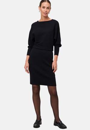 Femme portant un haut en tricot noir à manches longues et une jupe au genou, des collants noirs transparents et des mocassins noirs, se tenant devant un fond blanc.