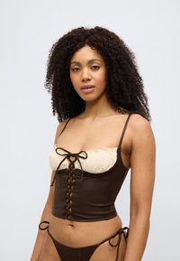 Καφέ κορσέ τοπ με λαιμόκοψη από ανοιχτό κρεμ βολάν, λεπτομέρεια δέσιμου στο μπροστινό μέρος και λεπτές τιράντες· λεία υφή με εφαρμοστή σχεδίαση.