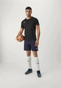 Mann i svart Puma-trøye, marineblå shorts, hvite sokker og fotballsko som holder en oransje og svart fotball mot en enkel bakgrunn.