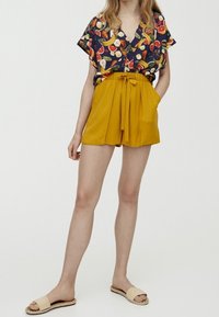 Short-sleeve, v-neck top with a fruit print in various colors, paired with high-waisted yellow shorts with a tie waist. Beige flat sandals.