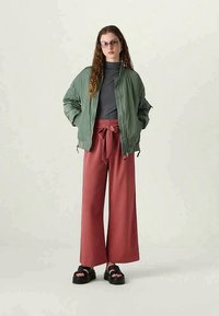 Olive green bomber jacket, gray t-shirt, wide-leg rust pants with a tied waist, and black sandals with thick soles. Simple design and relaxed fit.