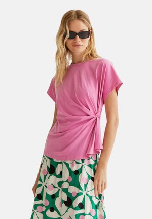 Mujer rubia con gafas de sol negras, una camiseta rosa de manga corta con nudo lateral y una falda verde con un patrón floral abstracto en blanco, rosa y negro.