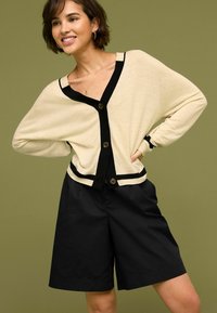 Gilet crème avec accents et boutons noirs, associé à un short noir. Le gilet présente une coupe décontractée et des poignets côtelés.