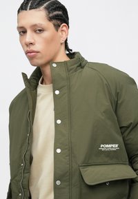 Casaco verde-oliva com fecho de correr e botões de pressão, com um bolso no peito e marca na frente. Textura suave, material leve.