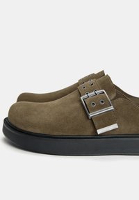 Olijfgroene suède schoenen met een ronde neus, zilveren gespdetail en een zwarte rubberen zool. Gladde textuur en minimalistisch ontwerp.
