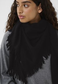 Sort tørklæde med frynser, lavet af blødt materiale, draperet over skuldrene, båret over en grå sweater.