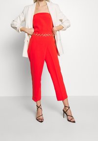 Combinaison rouge sans bretelles avec jambes fuselées et ceinture-chaîne argentée, associée à un blazer blanc et des talons hauts noirs à lanières.