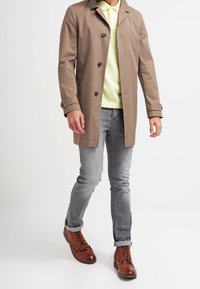Světlé hnědé trenčkot s černými knoflíky, nošený přes žluté polo tričko. V kombinaci se šedými džíny a hnědými šněrovacími botami.