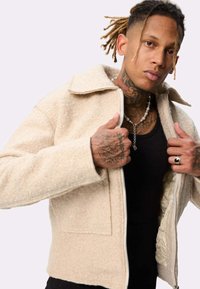 Veste beige zippée en tissu texturé avec un col pointu et deux poches avant. L'intérieur est doublé de matelassé.