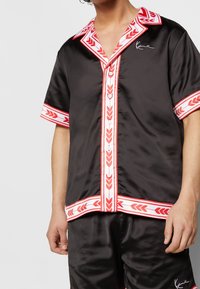 Man wearing black satin short-sleeve shirt and shorts with red and white patterned trim and white buttons, standing against a plain background.