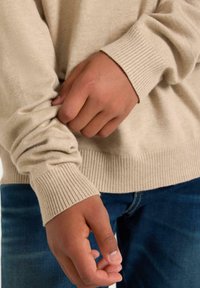 Beige trui met geribde manchetten en zoom; gladde textuur; handen die stof bij de taille vasthouden, gedragen met blauwe jeans.