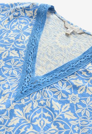 V-hals tunika med et blåt og hvidt blomstermønster, med en tekstureret blå crochetedetalje langs halsudskæringen. Blødt, let stof.