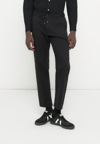 Pantalones con cintura elástica negra y cordón, con un diseño de pierna recta. Combinados con zapatillas negras con detalles blancos, sobre un fondo liso.