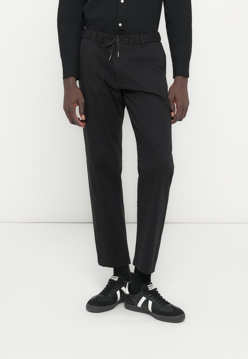 Pantalones con cintura elástica negra y cordón, con un diseño de pierna recta. Combinados con zapatillas negras con detalles blancos, sobre un fondo liso.