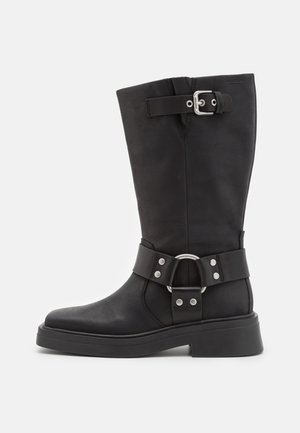 Bottes noires en cuir haute jusqu'au genou avec une finition mate, dotées d'une large sangle avec une boucle circulaire argentée et des détails à rivets. Semelle épaisse.