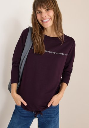 Maglietta a maniche lunghe viola con scollatura arrotondata, con testo bianco e orlo regolabile. Abbinata a Jeans blu.