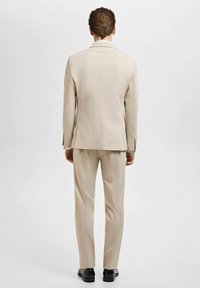Beige Anzug mit taillierter Passform, bestehend aus einem einreihigen Sakko und passenden Hosen, kombiniert mit schwarzen Schuhen. Glatte Textur.