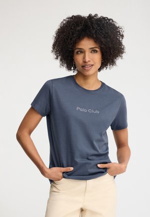 Frau mit lockigem Haar, die ein dunkelblaues "Polo Club"-T-Shirt und beige Hosen trägt, Hände in den Taschen, schaut leicht zur Seite.