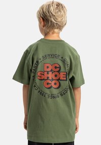 Olivově zelené bavlněné tričko s krátkým rukávem a kulatým výstřihem. Velká grafika na zadní straně zobrazuje "DC SHOE CO" a text v černé a oranžové barvě.