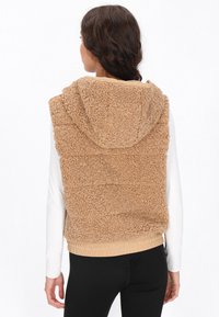 Beige Fleece-Weste mit Kapuze, die eine strukturierte Oberfläche, einen elastischen Bund und eine einfarbige beige Fütterung aufweist.