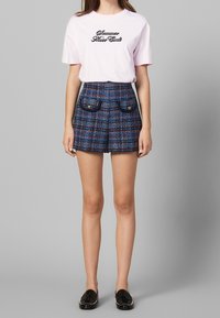 Femme portant un t-shirt rose avec le texte "L'été ne finit jamais", un short à carreaux bleu taille haute avec des poches à bouton, et des mocassins noirs à franges.