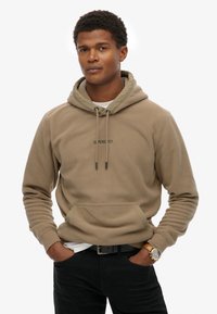 Beige fleece hoodie met een kangoeroezak, verstelbare trekkoordkap en groen logo. Gepaard met zwarte jeans en een horloge.
