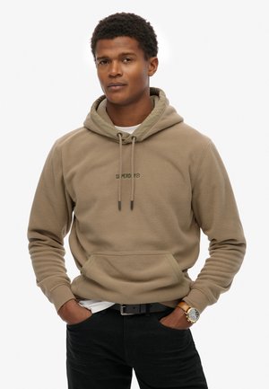 Beige fleece hoodie met een kangoeroezak, verstelbare trekkoordkap en groen logo. Gepaard met zwarte jeans en een horloge.