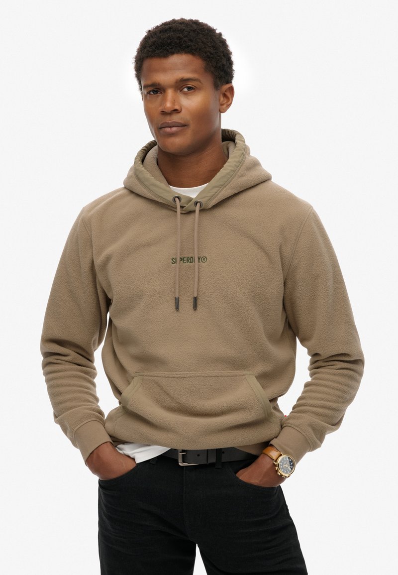 Beige fleece hoodie met een kangoeroezak, verstelbare trekkoordkap en groen logo. Gepaard met zwarte jeans en een horloge.