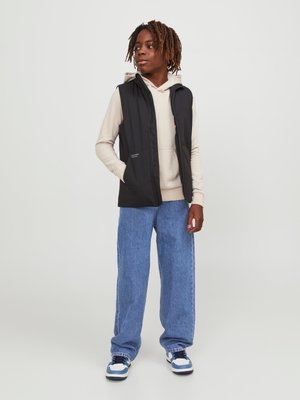 Jeune avec des dreadlocks portant un sweat à capuche beige, une veste sans manches noire, un jean bleu ample et des baskets blanches et bleues, debout devant un fond uni.