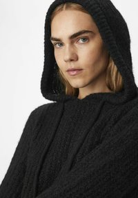 Zwarte hoodie gemaakt van textuurstof, met een ontspannen ontwerp en zichtbare stiksels langs de capuchon en naden.