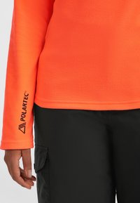 Sudadera de forro polar naranja brillante con una superficie texturizada. Presenta un logo de Polartec negro y un detalle en los puños. Combinada con pantalones cargo negros.