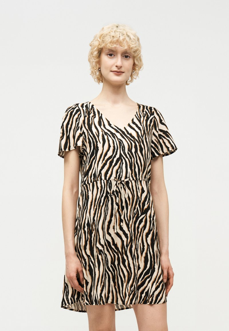 Ξανθιά γυναίκα με κοντομάνικο φόρεμα με V λαιμόκοψη και ρίγες τίγρης, με ζώνη στη μέση, που στέκεται μπροστά σε απλό ανοιχτόφθορο φόντο.