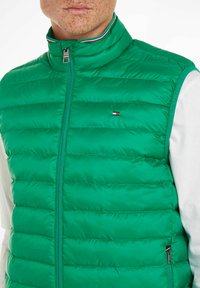 Grüne Steppweste mit horizontalem Steppdesign, hohem Kragen mit roten, weißen und blauen Akzenten sowie einem Reißverschluss vorne. Kleines Logodetail sichtbar.