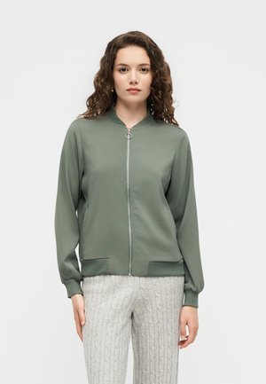Femme aux cheveux bouclés portant une veste verte à fermeture éclair et un pantalon gris clair en maille torsadée, debout devant un fond uni clair.