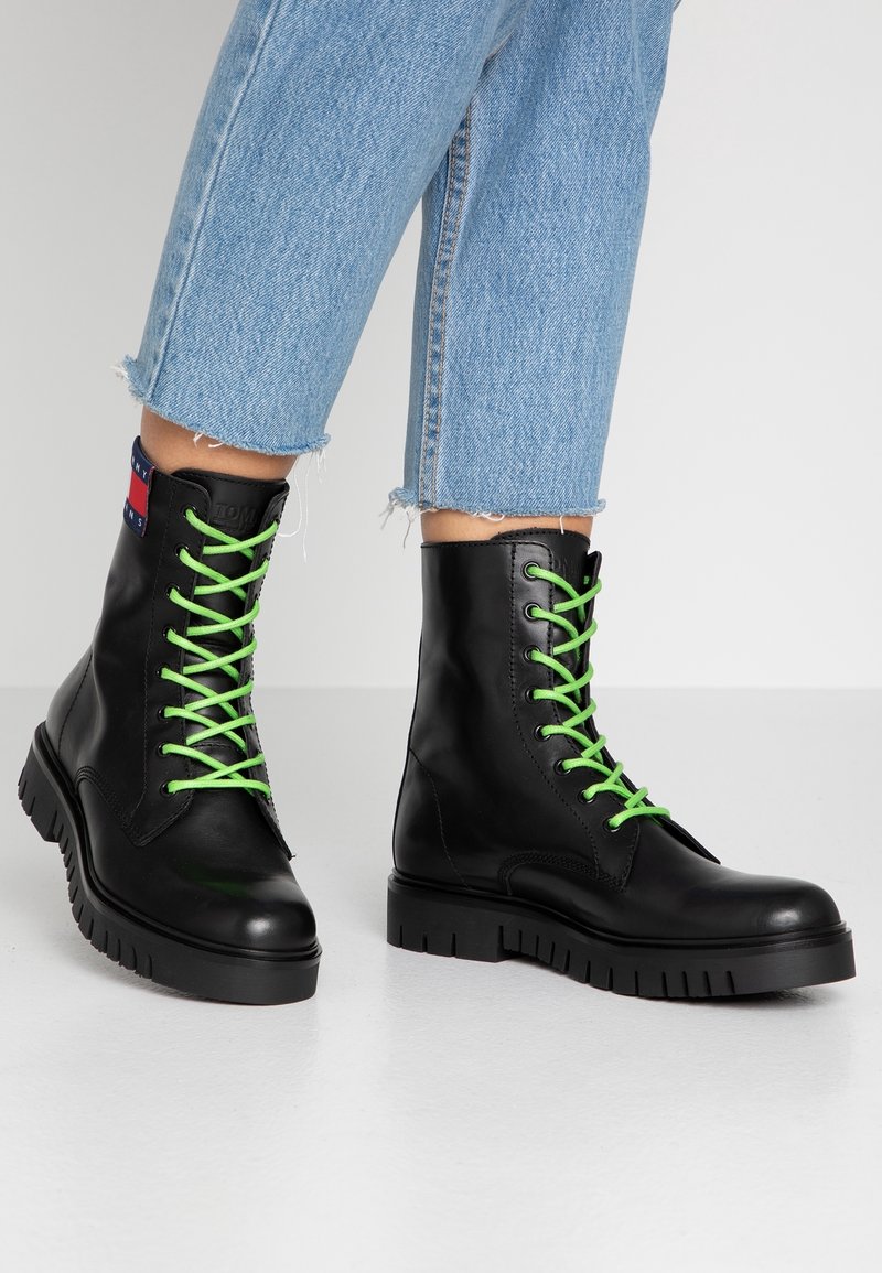 Bottes de combat en cuir noir avec des lacets vert vif portées avec un jean en denim bleu clair court, sur un fond gris uni.