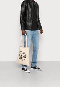 Béžová taška přes rameno s černým grafickým designem, z plátna, s dlouhými uchy; nošena s černou koženou bundou, černým tričkem, modrými džíny a teniskami.