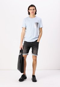 T-shirt bleu clair à manches courtes avec un design de poche grise, associé à un short en denim foncé et des chaussures noires. Le mannequin tient un sac fourre-tout noir.