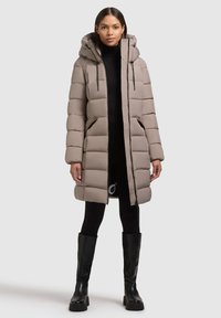 Beige színű puffertakó kapucnival, tűzéssel ellátott panelek, elülső zsebek és cipzár található rajta. A modellen fekete leggings és térd alatt érő csizma van.
