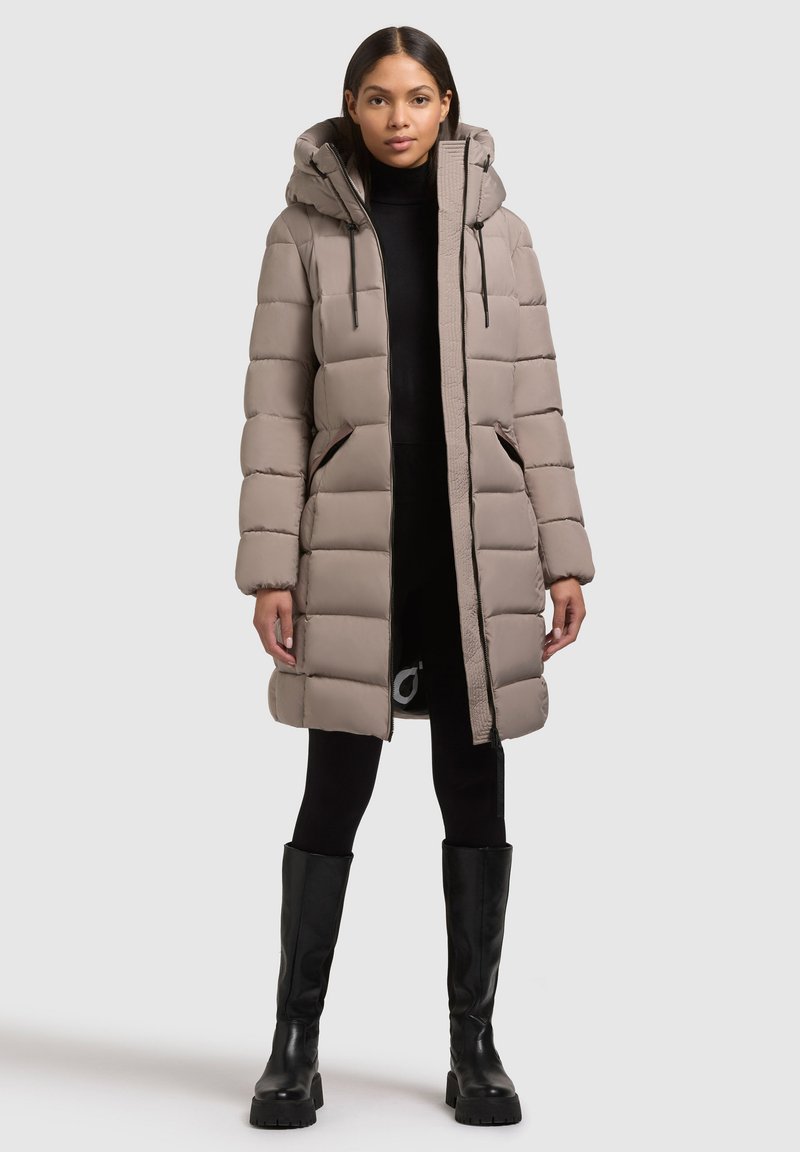 Beige színű puffertakó kapucnival, tűzéssel ellátott panelek, elülső zsebek és cipzár található rajta. A modellen fekete leggings és térd alatt érő csizma van.