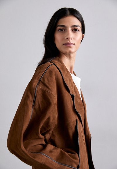 Giacca in suede marrone con una vestibilità rilassata, caratterizzata da piping nero lungo il colletto e la parte frontale. Texture liscia, forma angolare e maniche larghe.