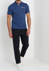 Polo bleu avec un col court et un logo "SUNSET" brodé, accompagné d'un pantalon foncé et de baskets blanches.