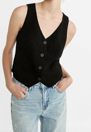 Vrouw die een mouwloos zwart vest met knopen draagt over lichtblauwe high-waisted jeans met handen in de zakken, tegen een witte achtergrond.