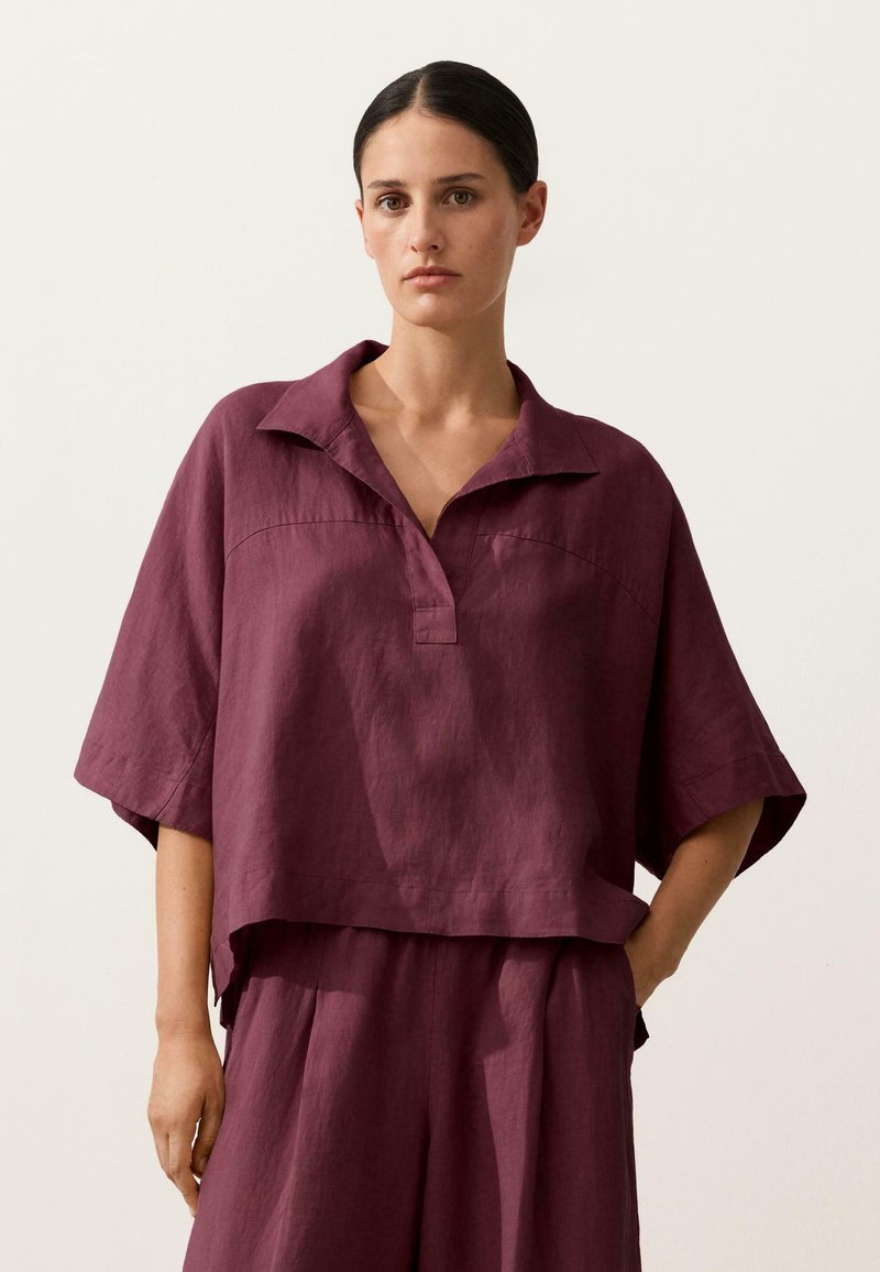 Femme aux cheveux foncés portant un haut prune à manches courtes amples avec col et un pantalon taille haute large assorti, debout devant un fond uni.