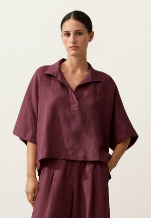 Sötét hajú nő laza, rövid ujjú, galléros szilvalila felsőben és hozzá illő magasderekú, bő szárú nadrágban, egyszínű háttér előtt áll.