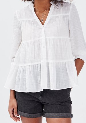 Femme portant un chemisier blanc à boutons à volants avec des manches trois-quarts et un short en jean noir à ourlet roulé, accessoirisé avec des colliers superposés.